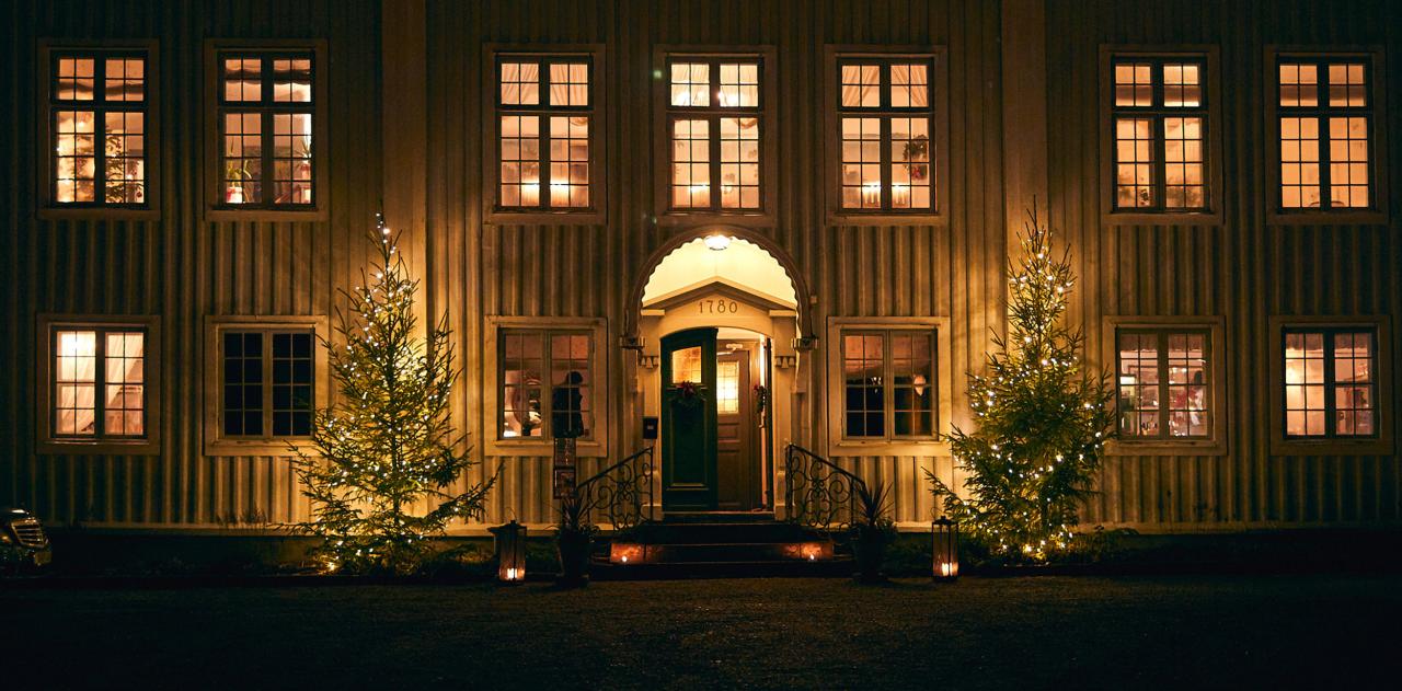 herrgård utifrån på kvällen, lyser i alla fönster, julgranar framför ingång